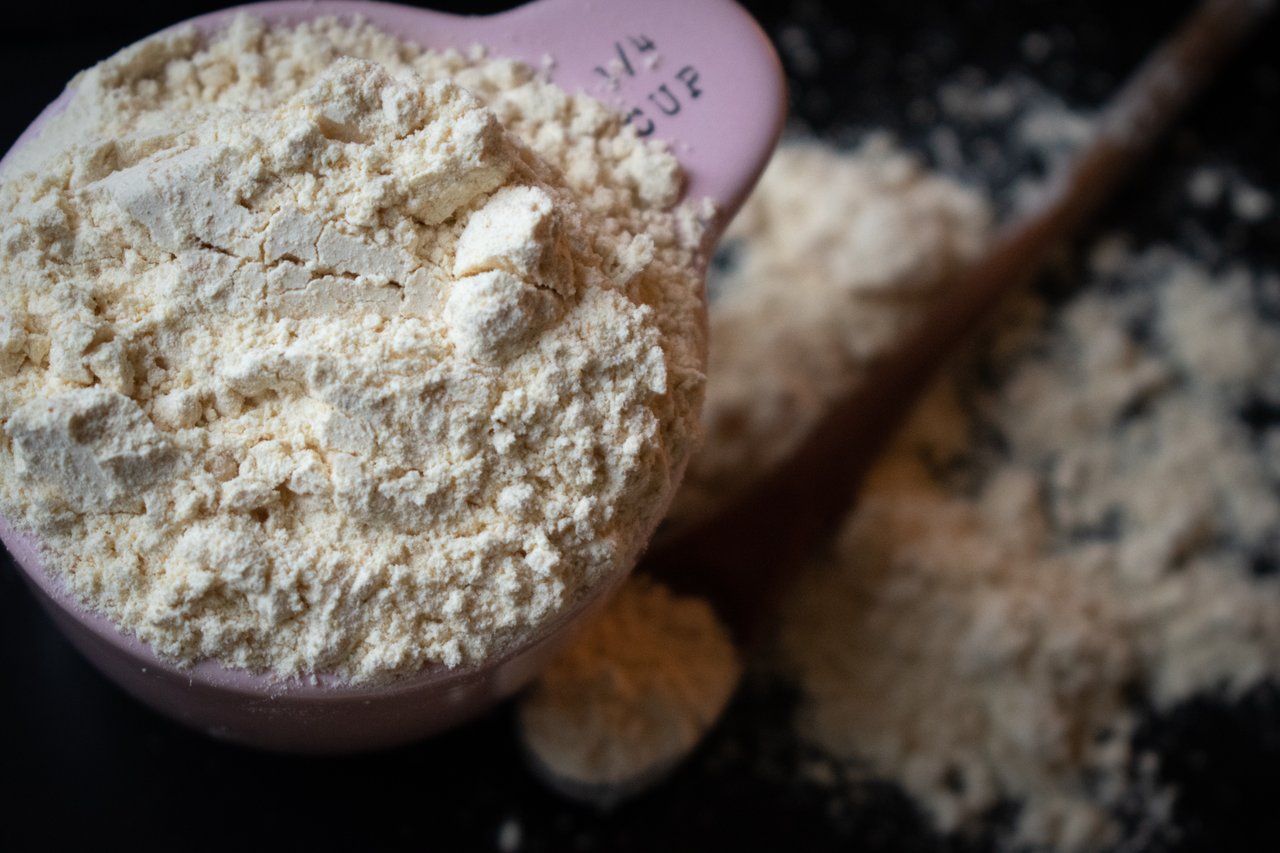 about-01 A filled measuring cup of flour spills onto a dark surface in a shallow depth of field.